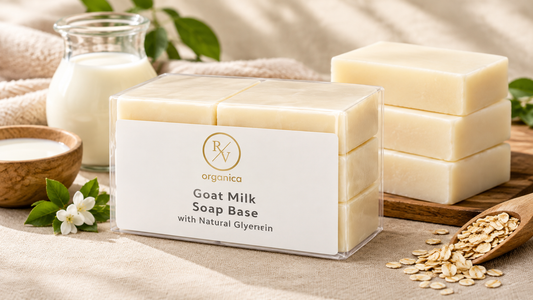 RV Organica Goat Milk Soap Base with Natural Glycerin in a clear pack beside stacked creamy soap bars, a jug of milk, oats and jasmine flowers on beige linen