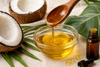 Cold-pressed coconut oil being poured from a wooden spoon into a glass bowl, with fresh coconut halves, tropical leaves, and an essential oil bottle on a linen background