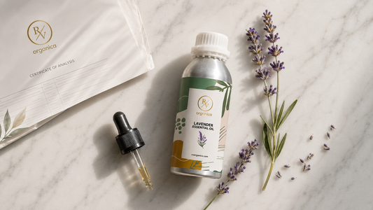 Flat lay product photography of RV Organica Lavender Essential Oil aluminum bottle with dropper, Certificate of Analysis (COA) document, fresh lavender sprigs, and scattered lavender buds on a white marble surface under soft natural light