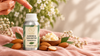 RV Organica Almond & Shea Butter Fragrance Oil bottle with almonds and shea butter on blush pink linen, featuring a golden oil drop in a soft natural beauty setting.