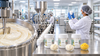 Quality technician in a hairnet and gloves inspecting a jar in a clean body butter manufacturing facility, with a steel mixing vessel, filling line and RV Organica Shea, Cocoa and Mango butter jars