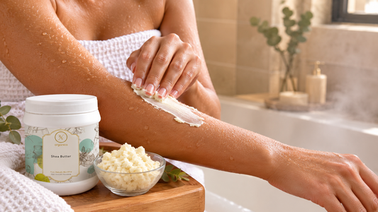 Woman wrapped in a towel smoothing shea butter onto her damp forearm after a bath, with an RV Organica Shea Butter jar and a bowl of shea butter on a wooden tray