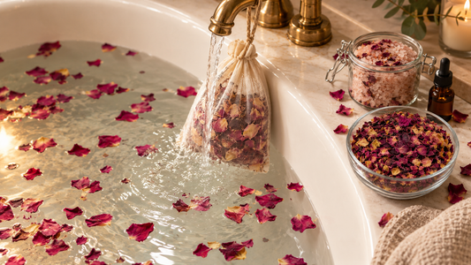 Dried rose petals in a muslin bag under a running bath tap with petals floating in the water, a jar of pink bath salt and a dropper bottle of rose oil
