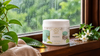 RV Organica Shea Butter jar on a wet wooden windowsill with raindrops on the glass, green foliage outside, jasmine flowers and a towel