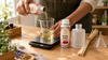 Hands pouring RV Organica Royal Rose fragrance oil into a glass beaker on a digital scale, next to a frosted reed diffuser bottle, a bundle of rattan reeds and a small funnel on a wooden workbench
