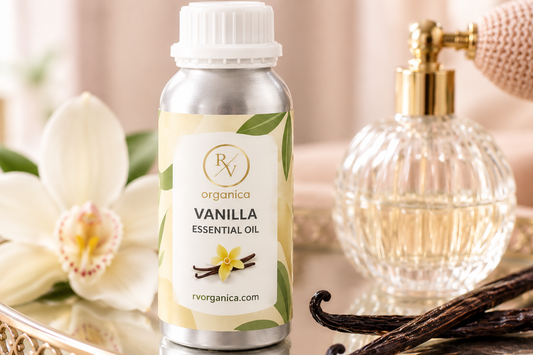 RV Organica Vanilla Essential Oil bottle on a mirrored vanity tray beside a vintage glass perfume atomizer, a white orchid and cured vanilla pods