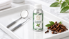 RV Organica Clove Leaf Essential Oil in a silver bottle with a green botanical label, displayed on a white tray alongside stainless steel dental tools and dried clove buds.