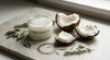 A glass jar of coconut oil beside cracked coconut halves, dried herbs, and strands of healthy dark hair on a stone surface in soft natural light.