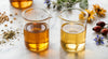 Two glass beakers of golden carrier oil in different shades placed side by side on a marble surface with botanical seeds scattered in the background.
