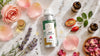 Luxury flat lay of RV Organica Rose Essential Oil aluminum bottle on white marble with fresh pink rose petals, lavender sprigs, amber essential oil bottles, rosehip fruit, and golden serum drops in soft natural morning light.